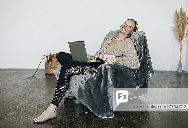 Smiling woman holding coffee cup sitting with laptop on armchair at home