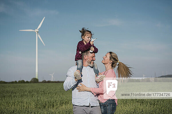 Smiling man carrying daughter on shoulder holding wind turbine model standing by woman at field