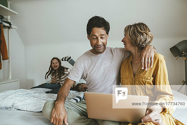 Mann mit Laptop sitzt neben Frau und schaut Tochter und Sohn zu  die zu Hause im Bett spielen