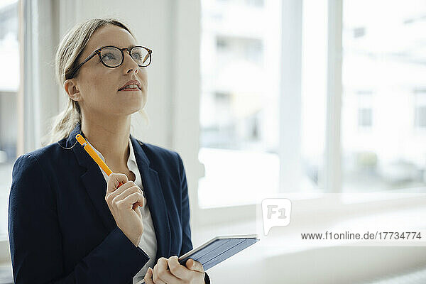 Thoughtful businesswoman holding tablet PC looking up in office