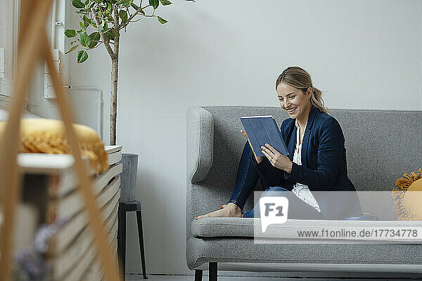 Happy businesswoman looking at tablet PC sitting on sofa in office