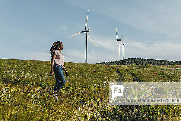 Smiling young woman walking on meadow in front of wind turbine