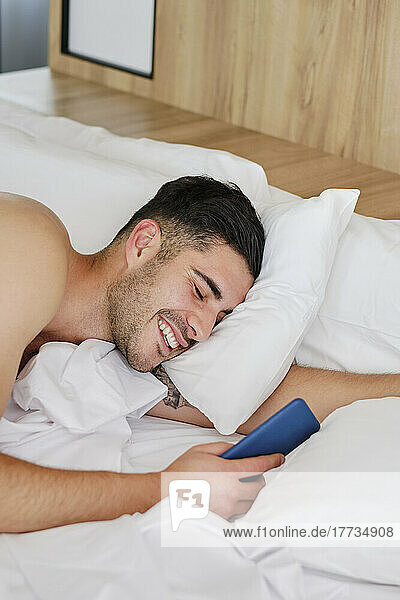 Happy man holding mobile phone waking up in bed at home