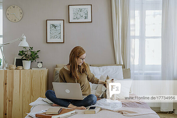 Smiling woman sitting with laptop stroking pet dog on bed at home