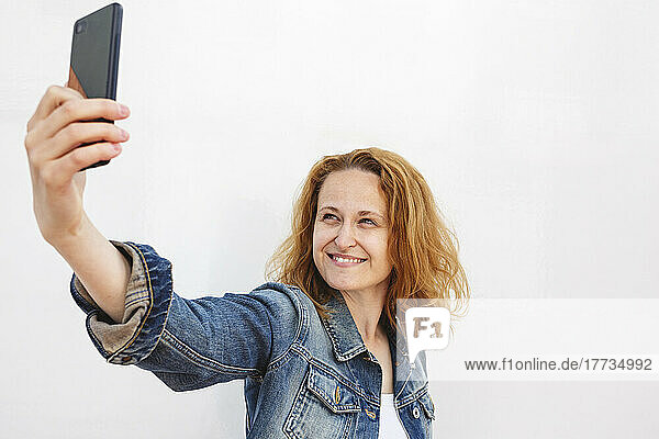 Smiling woman talking selfie through smart phone against white background