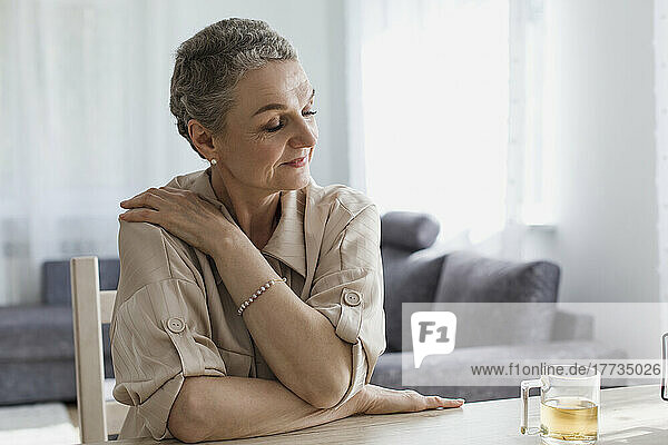 Mature woman sitting at table at home with closed eyes hugging herself