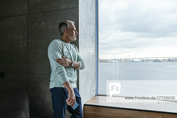 Thoughtful man holding mobile phone looking through window at home