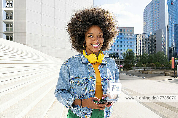 Happy young woman wearing wireless headphones standing with smart phone by steps on sunny day