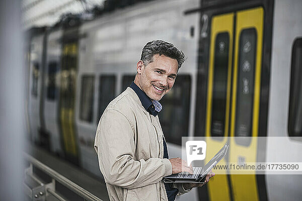Happy businessman with tablet PC standing in front of train at railroad station