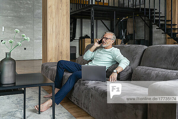 Businessman talking on smart phone sitting with laptop on sofa at home