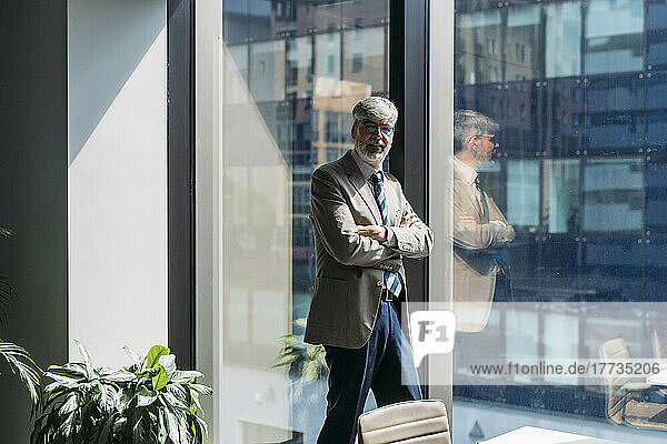 Businessman with arms crossed standing by glass window at office