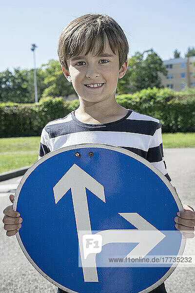 Smiling boy holding road sign board with arrow symbol on sunny day