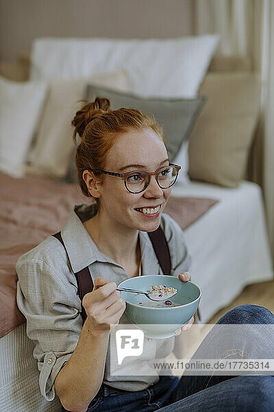 Happy woman wearing eyeglasses sitting with bowl of food in front of bed at home