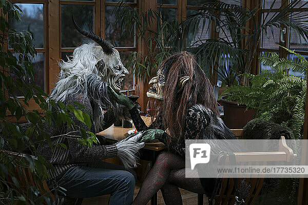 Krampus and witch with wineglass holding hands sitting at table