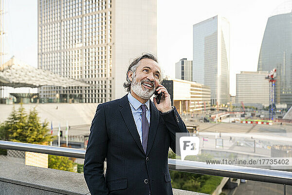 Happy mature businessman talking on smart phone standing by railing