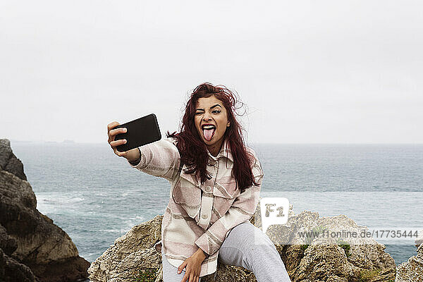 Woman sticking out tongue sitting on rock taking selfie through mobile phone