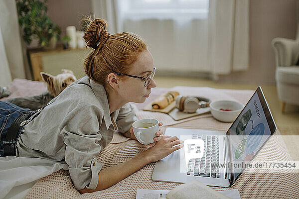 Frau mit Kaffeetasse und Laptop liegt zu Hause im Bett