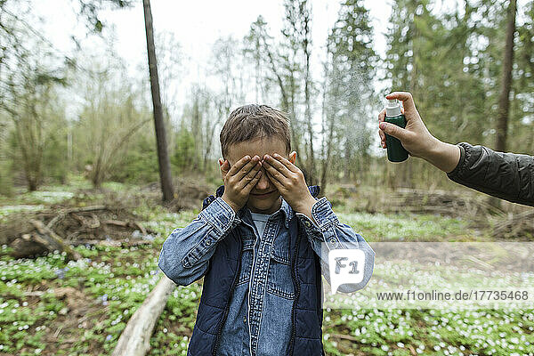 Mother spraying insect repellant on son covering eyes in forest