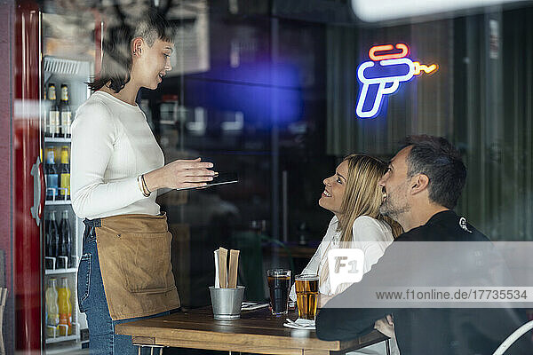 Waitress talking with customers sitting at table seen through glass