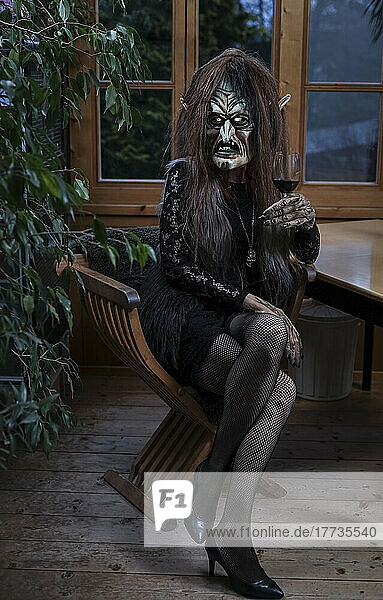 Frau im Hexenkostüm sitzt mit Weinglas auf Stuhl im Horrorhaus
