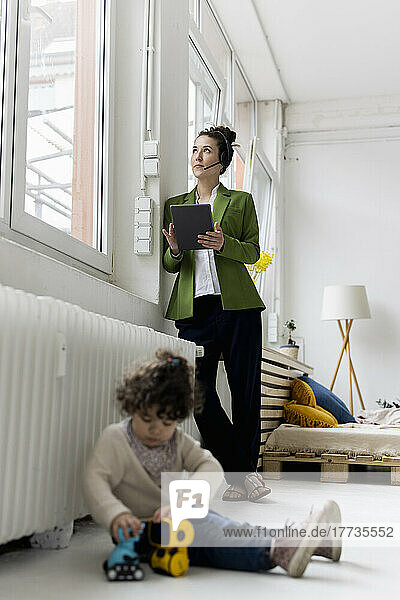 Young businesswoman standing at window working  whilde daughgter is playing on floor