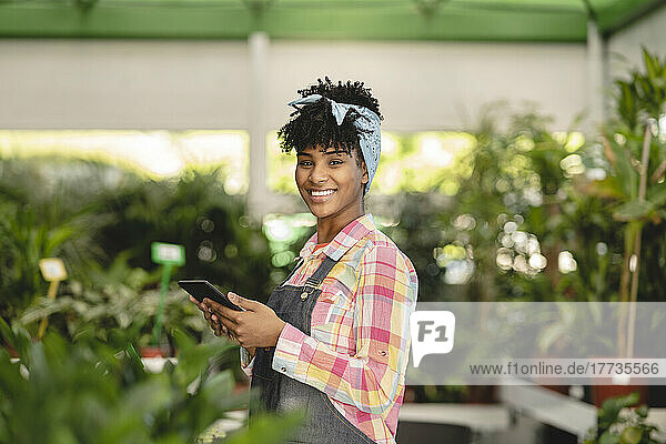 Happy gardener holding tablet PC standing at plant nursery