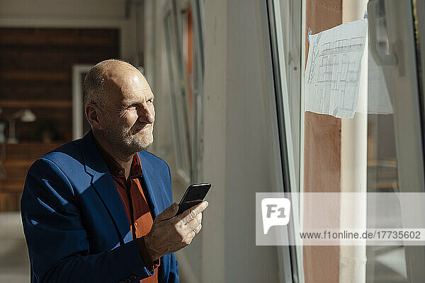 Geschäftsmann hält Mobiltelefon in der Hand und schaut auf den Bauplan am Fenster
