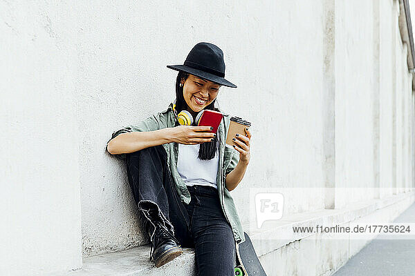 Happy woman with reusable coffee cup using smart phone sitting on wall