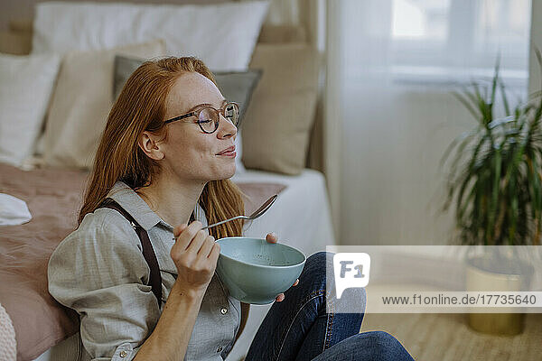 Smiling woman with eyes closed eating food sitting in front of bed at home