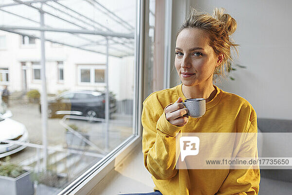 Frau hält Kaffeetasse und sitzt zu Hause auf der Fensterbank