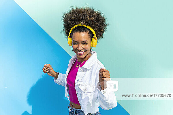 Happy woman listening music through wireless headphones dancing in front of blue wall