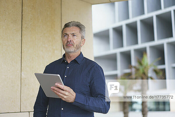 Thoughtful businessman holding tablet PC standing by wall