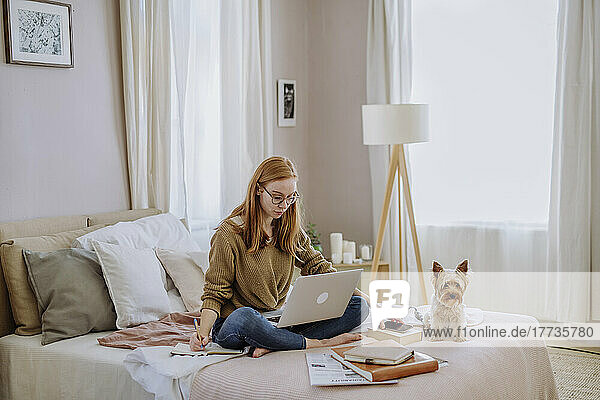 Woman writing in book sitting with laptop by pet dog on bed at home
