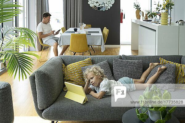 Girl watching tablet PC on sofa with father sitting on chair in living room at home