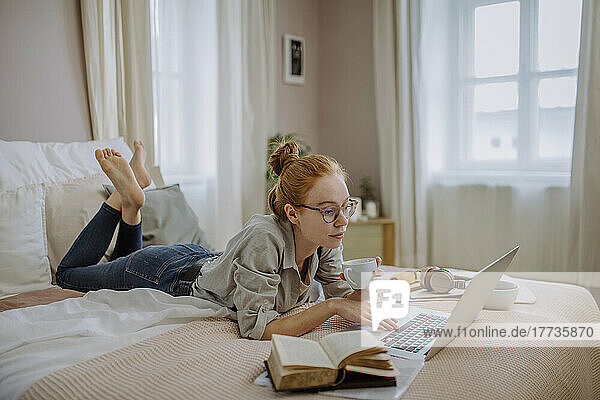 Woman wearing eyeglasses holding coffee cup using laptop lying on bed at home
