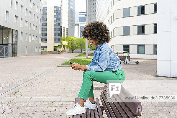 Smiling Afro woman text messaging through smart phone sitting on bench