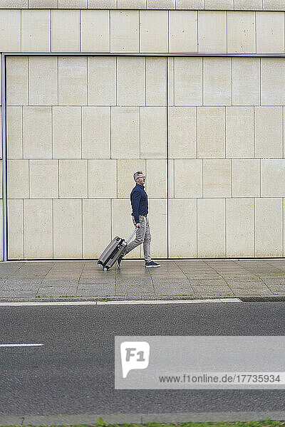 Businessman with suitcase walking on footpath by wall
