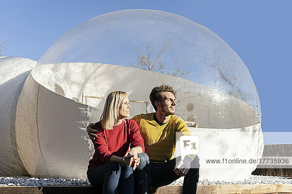 Junger Mann mit Freundin sitzt an einem sonnigen Tag vor einem transparenten Kuppelhotel