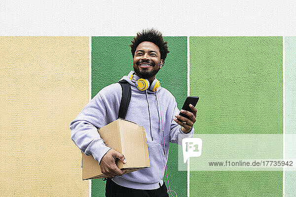 Lächelnder Lieferbote hält Karton und Mobiltelefon vor bunter Wand