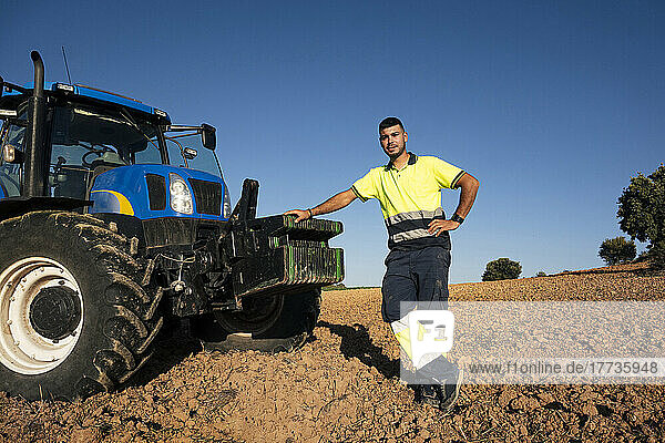 Young farmer with hand on hip standing by tractor on sunny day
