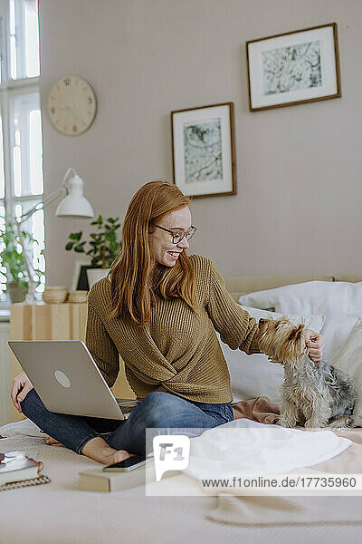 Happy woman sitting with laptop stroking pet dog on bed at home