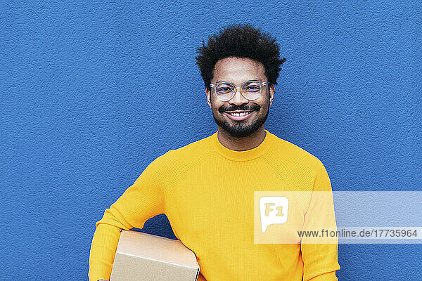 Smiling delivery man holding box in front of blue wall