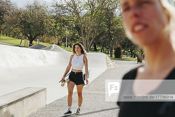 Smiling woman holding skateboard walking near friend at sports ramp on sunny day