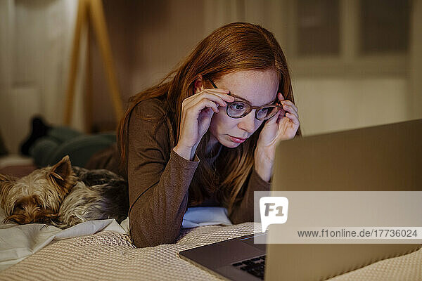 Woman wearing eyeglasses lying with laptop by pet dog on bed at home