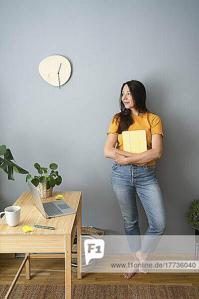 Smiling woman looking away holding diary in front of gray wall