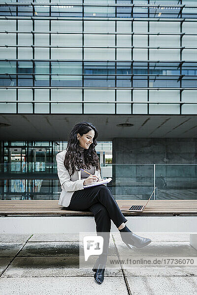 Smiling young businesswoman sitting on bench writing in book looking at laptop