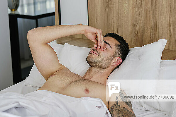 Shirtless man rubbing eyes lying in bed at home