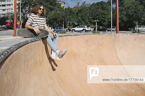 Young man with skateboard sitting at sports ramp on sunny day