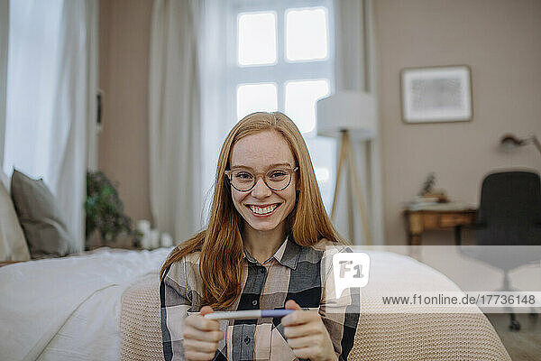 Glückliche rothaarige Frau mit Brille sitzt zu Hause mit Schwangerschaftstest vor dem Bett