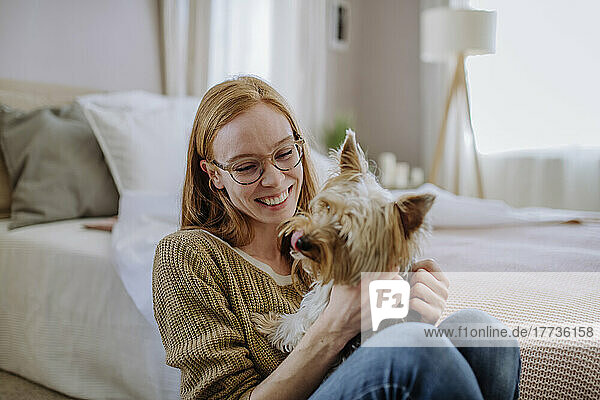Cheerful woman with pet dog sitting by bed at home